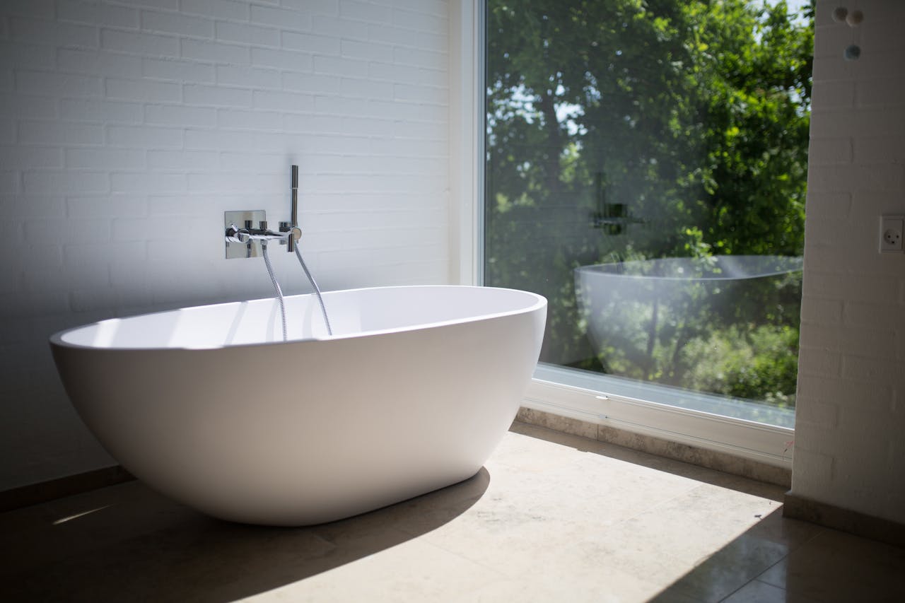 contact-img-01 Sleek white bathtub by large window in sunlit minimalist bathroom setting.